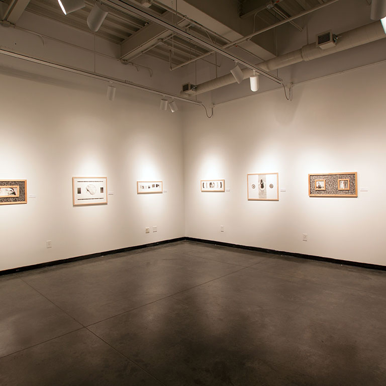 A gallery space with black floors, white walls, and frames hanging on the walls.
