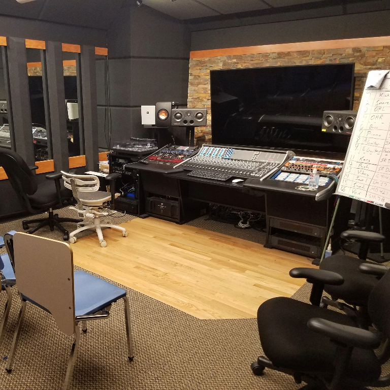 Color photograph of the CLEAR lab at Herron School of Art and Design. The space consists of a music recording console, speakers, a large screen, and lots of seating.