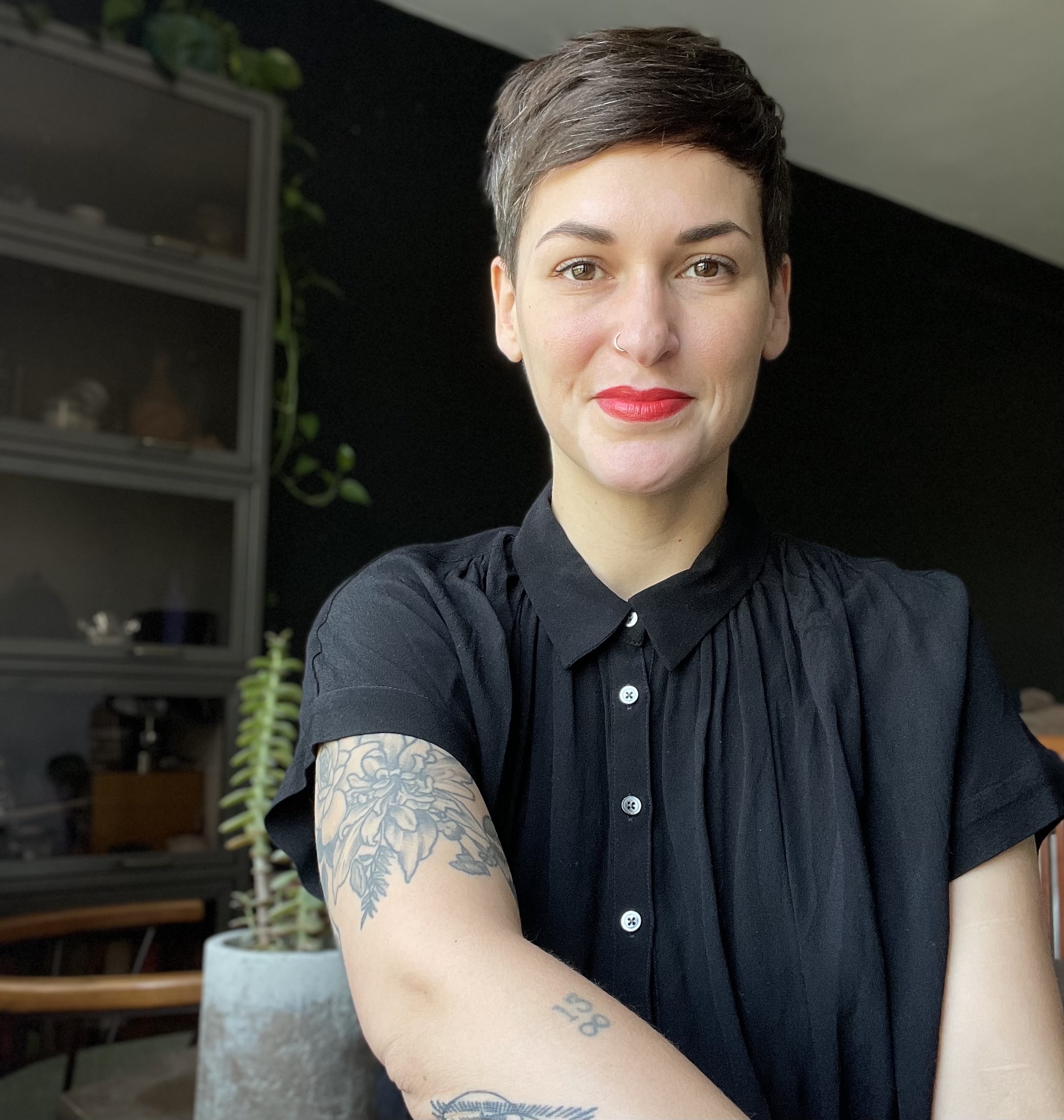 Color photograph of Kate Myers-Coffman seated in a black shirt with houseplants on a set of shelves in the background