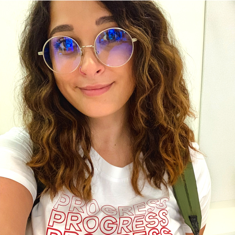 A profile picture of Natasha Chopra wearing round sunglasses and a t-shirt that reads "PROGRESS."