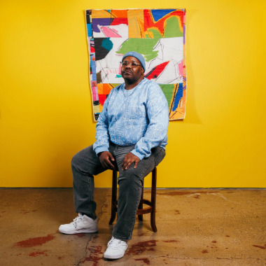 Artist Julian Jamaal Jones sitting on a wooden stool in front of a bright yellow gallery wall with one of his colorful quilts hanging behind him.