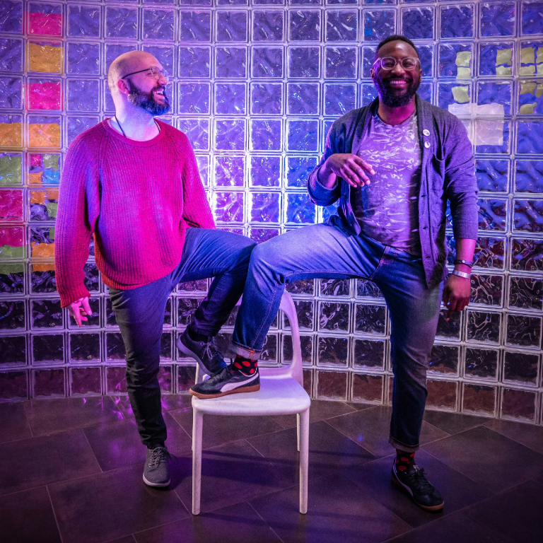 Color photograph with AMPL Labs cofounders Michael Runge on the left and Mayowa Tomori on the right smiling and posing in a purple-lit glassed in room