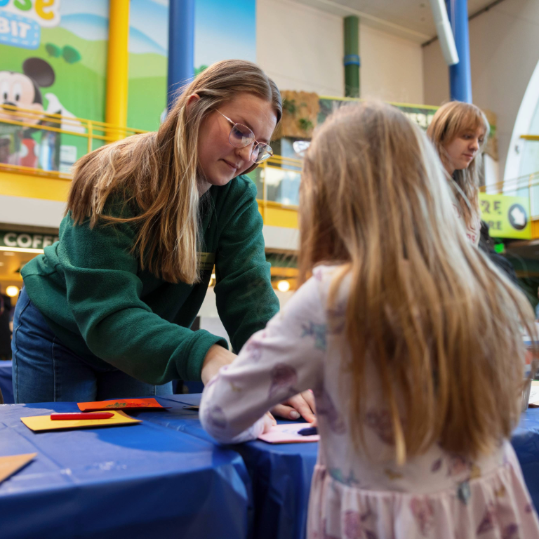 Herron student volunteer in glasses and a green sweatshirt holding down a piece of paper while a child with their back turned to the camera rubs a crayon across the paper