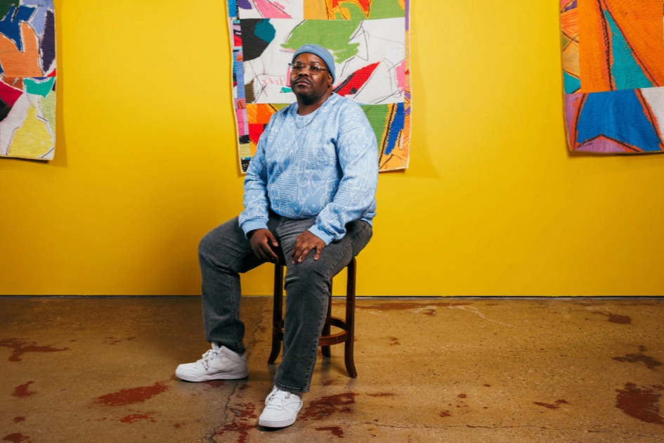 Artist Julian Jamaal Jones sitting on a wooden stool in front of a bright yellow gallery wall with three of his colorful quilts hanging on it.